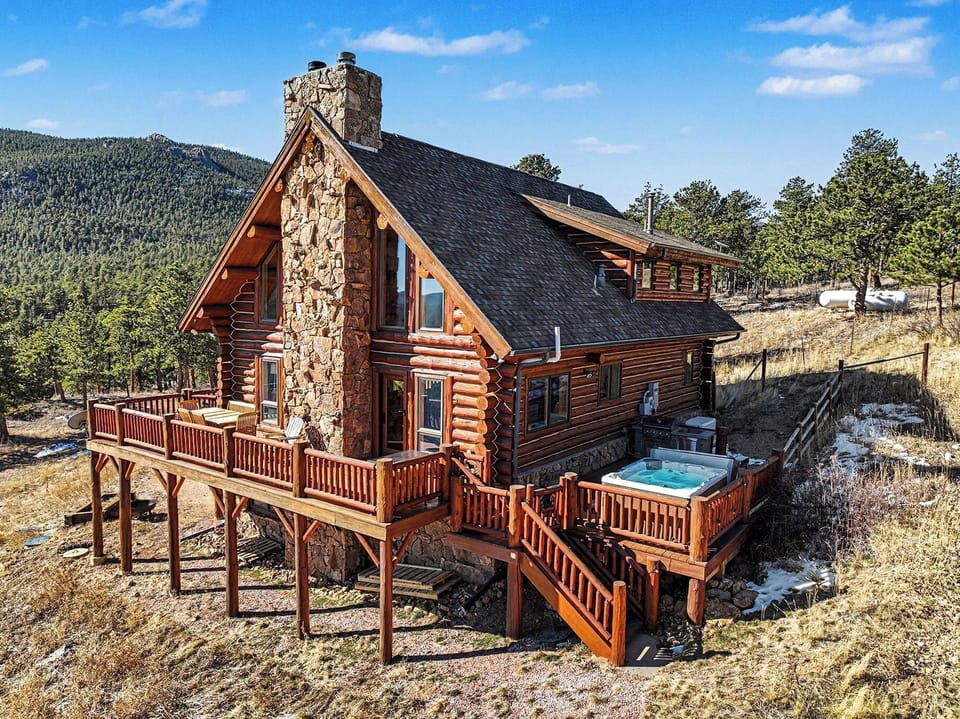 Rustic log cabin with stone chimney nestled in mountain landscape, featuring wraparound deck and hot tub amid natural surroundings.