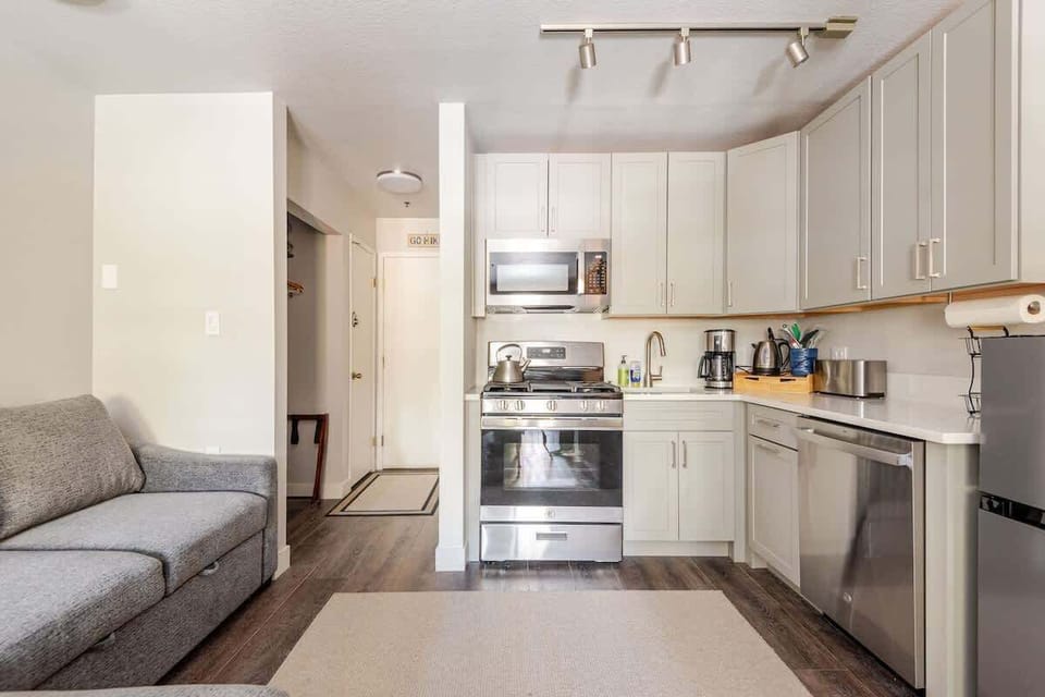 Cozy kitchen with gray sofa, stainless appliances, bright lighting, and inviting layout.