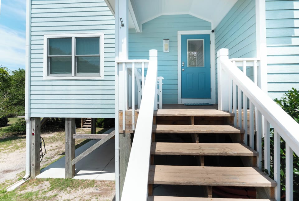 A charming coastal cottage painted in soothing aqua with white trim welcomes guests. The inviting front entrance features wooden stairs and white railings leading to your private retreat by the sea.