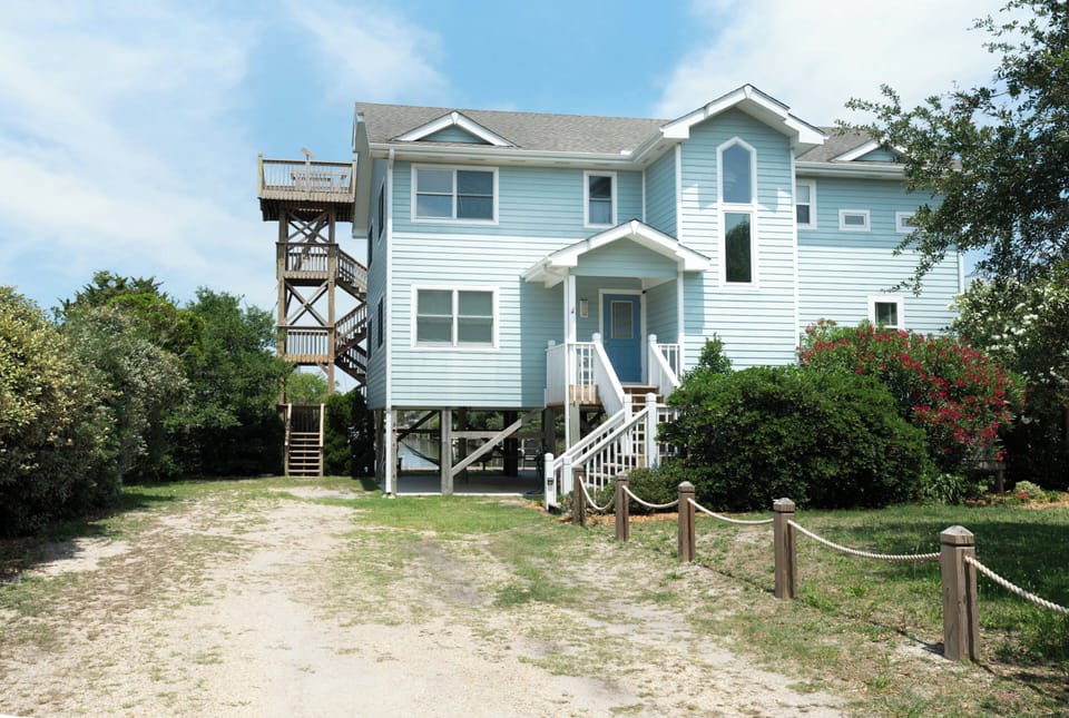 This charming coastal retreat features distinctive blue siding, elevated architecture, and a private observation tower, nestled among mature landscaping with nearby beach access via a sandy path.