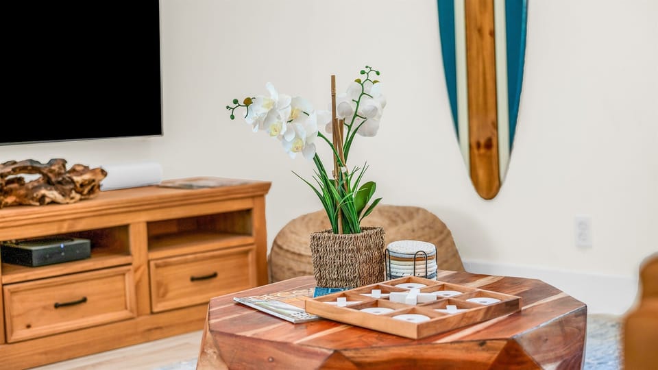 The bright and airy living room features a modern leather sectional, warm wood accents, and lush greenery views through large picture windows. Stylish touches like the surfboard décor and geometric coffee table create a relaxed coastal vibe.