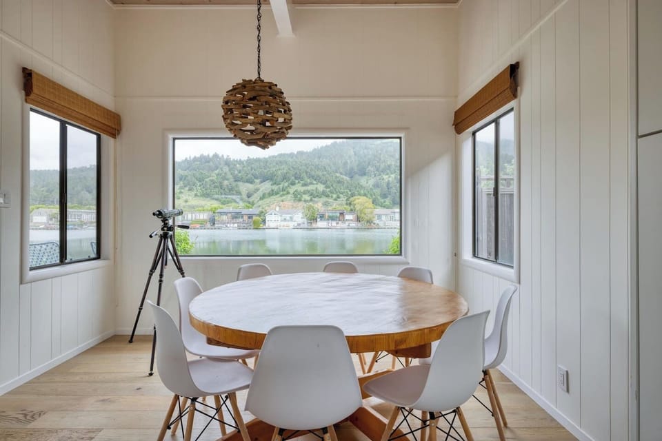 - Bright dining area with round wooden table and seating for eight guests  
- Stylish windows offer panoramic water and hillside views while dining  
- Unique pendant light adds charm to this airy, scenic gathering space  
