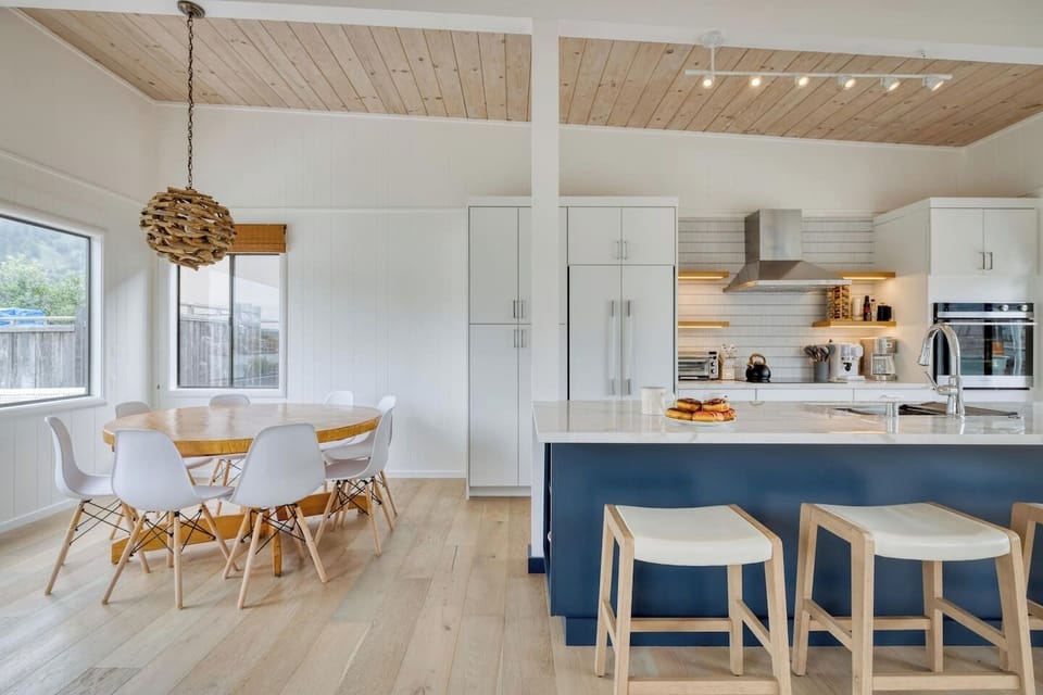 - Dining space with round table and modern chairs for shared meals  
- Kitchen features a navy island with bar seating for casual dining  
- Light wood flooring and ceiling create a warm, coastal inspired vibe  
