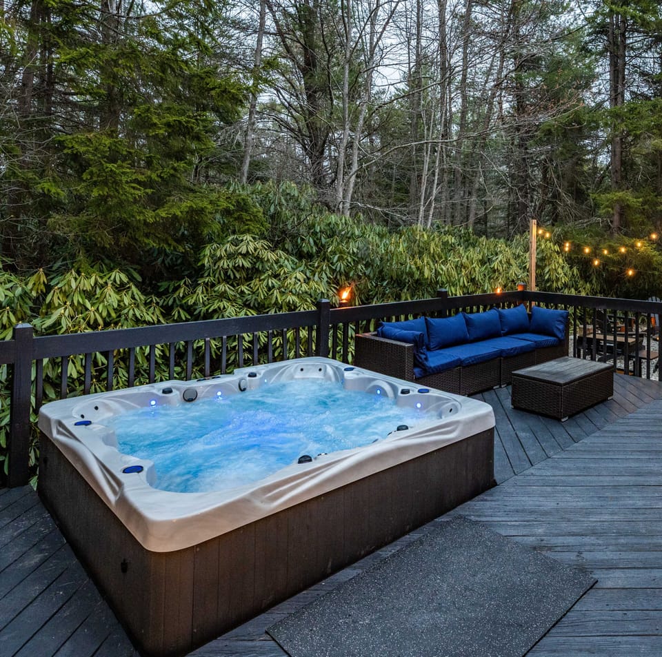 The deck is a private retreat with a bubbling hot tub, comfy outdoor seating, and string lights that lead to the fire pit area—perfect for unwinding under the trees.