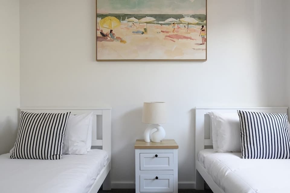 Rest easy in this bright, relaxing bedroom, perfect for a peaceful night's sleep. Featuring two comfortable beds with crisp white linens and stylish striped pillows, separated by a charming bedside table with a lamp.