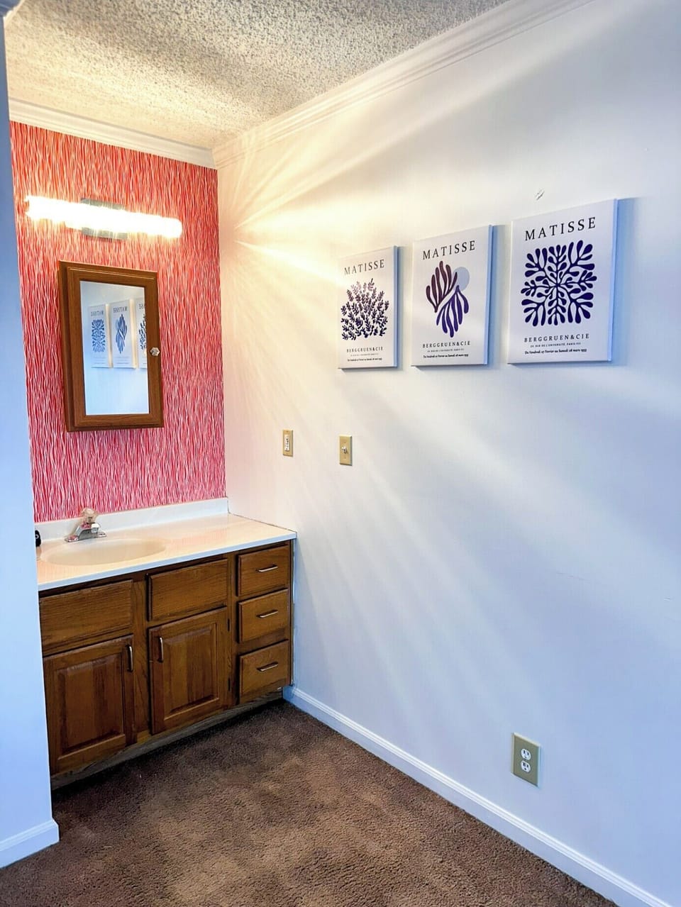 This bathroom offers a functional space with a wooden vanity and sink, complemented by a mirrored medicine cabinet and a bright light fixture. The combination of a vibrant red patterned accent wall and serene blue Matisse-inspired prints adds a unique artistic touch to the room.