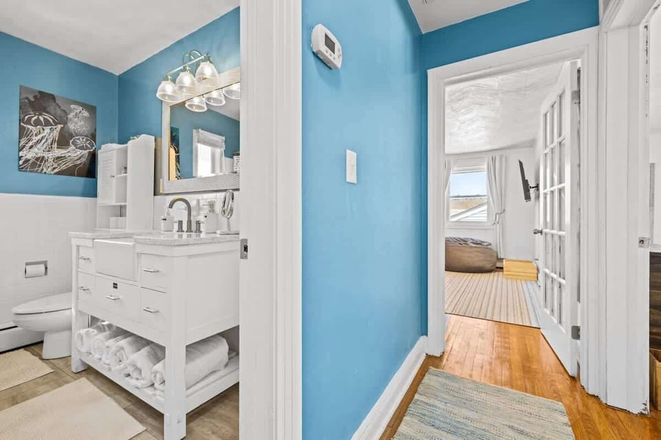 A white dresser and mirror form a lovely vanity corner. Blue walls and natural light make it bright and welcoming. 
