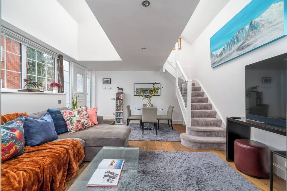 Bright and spacious open-plan living area with a plush sectional sofa, cozy rugs, and a dining table for six. A staircase leads to the upper level, while skylights and large windows flood the space with natural light. Stylish and welcoming throughout