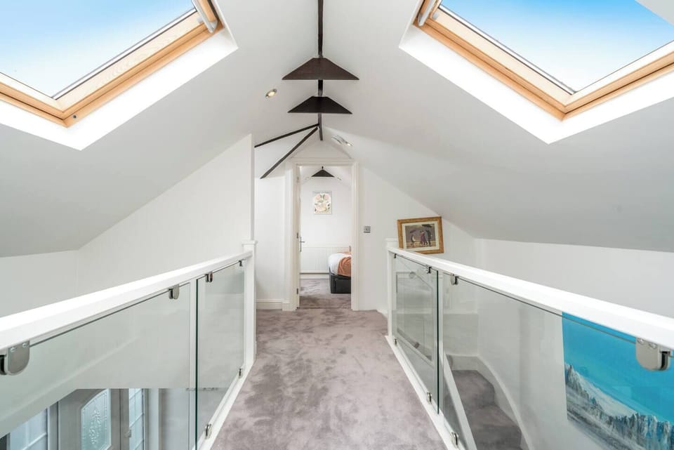 Stunning loft hallway with skylights, exposed beams, and plush carpeting. Glass balustrades add a modern touch while allowing natural light to flow through, leading to a cozy bedroom at the end of the walkway. Bright, airy, and architecturally unique