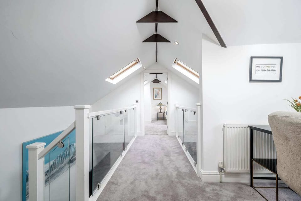 Bright loft hallway with skylights, vaulted ceilings, and exposed beams. Glass balustrades line the walkway, creating an open and airy feel. Soft carpeting adds comfort while leading to a cozy room at the end of the corridor.