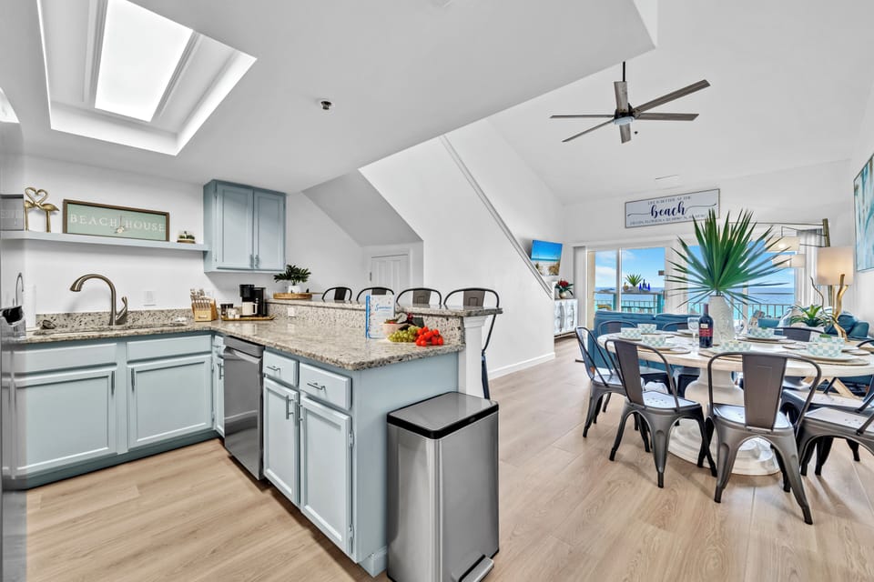 Bright ocean-view kitchen with granite counters, coastal blue cabinets, and dining space flooded with natural light from skylights.