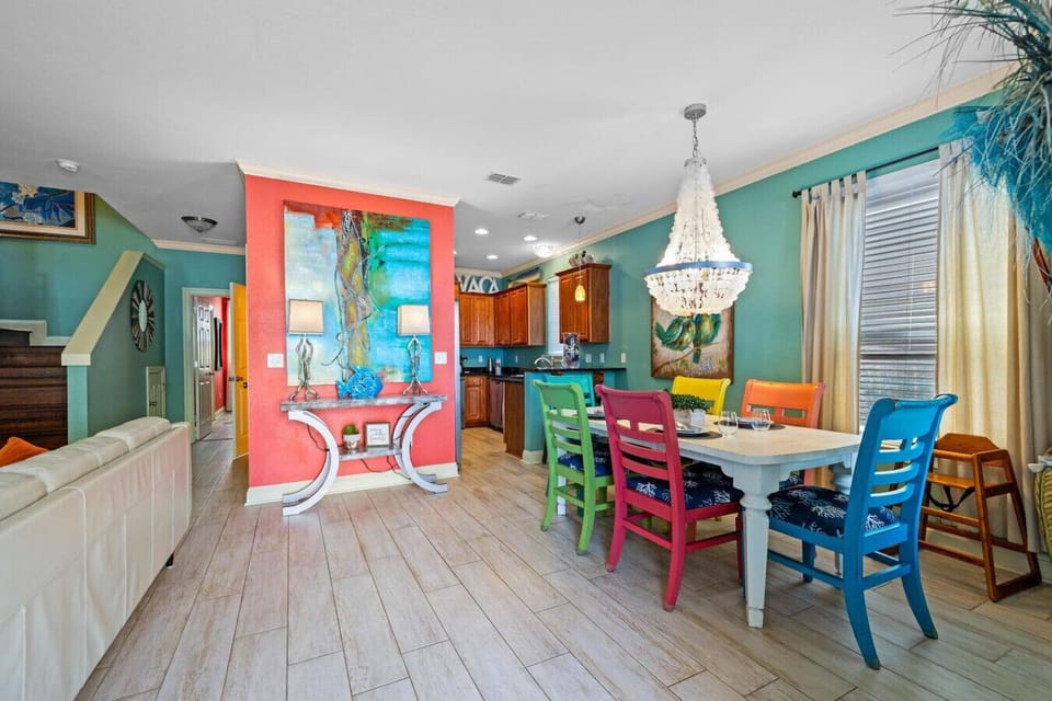 This open-concept living space is basically a masterclass in playful color—where turquoise walls, a coral accent kitchen, and rainbow dining chairs come together to prove that vacation homes don`t have to choose between stylish and fun.