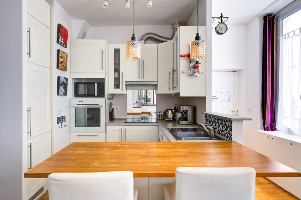 La pièce combine une cuisine ouverte et un espace repas chaleureux. Une table ronde en bois, des chaises modernes, un bar avec tabourets et des équipements complets. Une grande fenêtre créant une ambiance lumineuse et une pièce très conviviale.