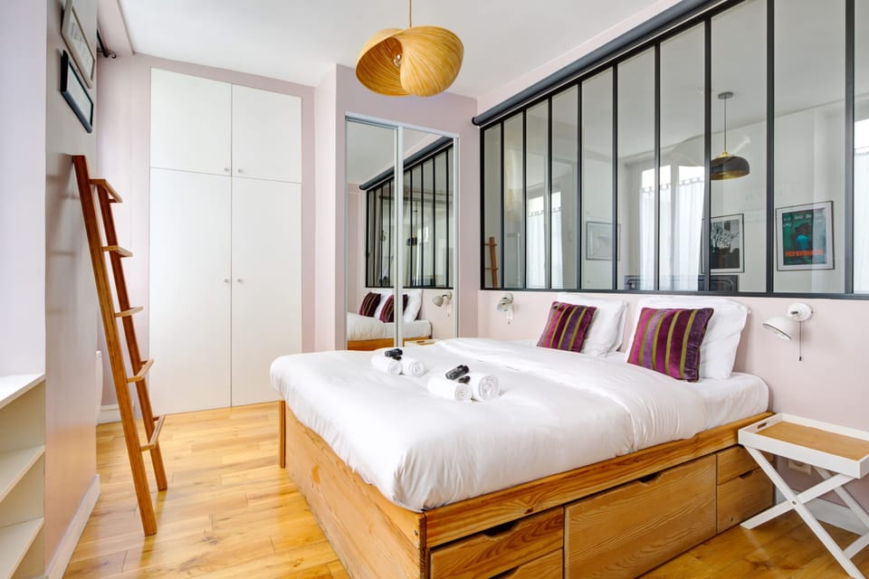 Chambre moderne et lumineuse avec lit double, coussins colorés et parquet en bois clair. Rangements intégrés sous le lit, armoire blanche et verrière noire apportant une touche industrielle élégante. Décoration soignée, confortable et chaleureux.