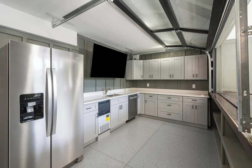 Outdoor kitchen next to pool with sink, smart tv, dishwasher, refrigerator, icemaker and propane grill.  Glass garage door opens to pool.