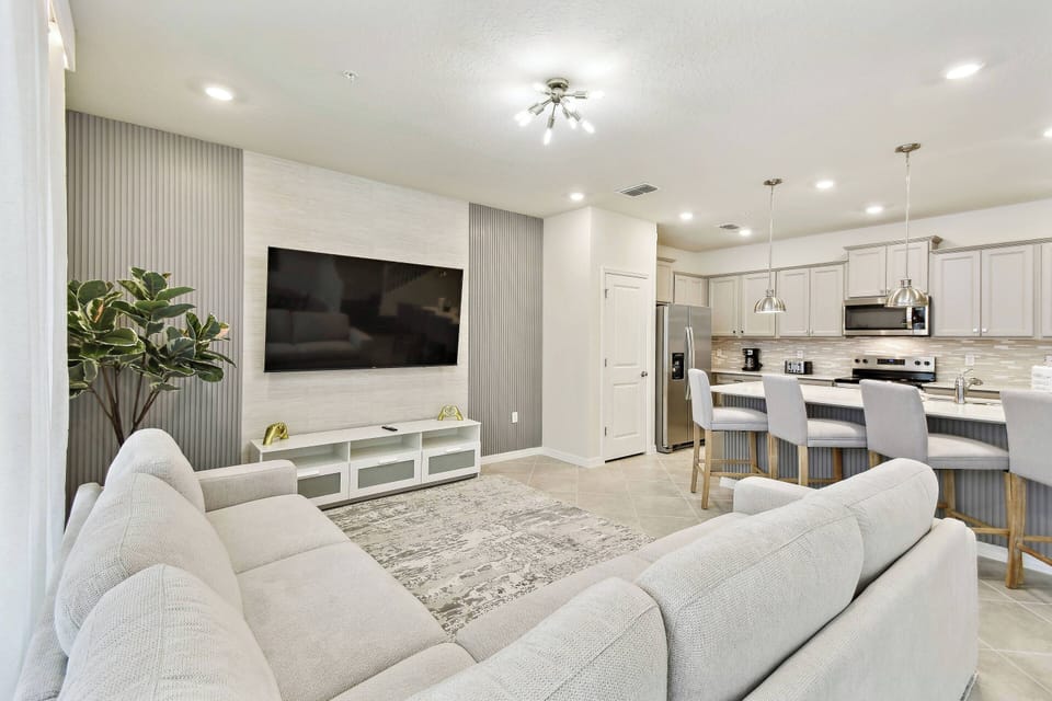 This stylish and spacious living area boasts a plush light-colored sofa perfect for relaxation, along with a large flat-screen TV on a sleek white media console, all seamlessly connecting to a modern kitchen ideal for socializing.