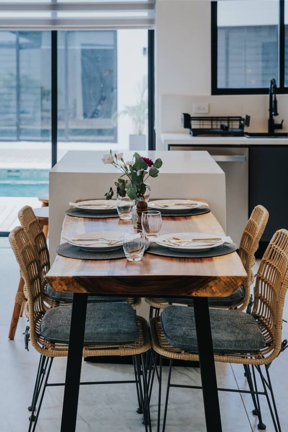 A beautifully set dining table for four, with two bar stools at the counter for a total of six seats. The modern kitchen and pool view create a fresh, open atmosphere, perfect for enjoying meals and entertaining.