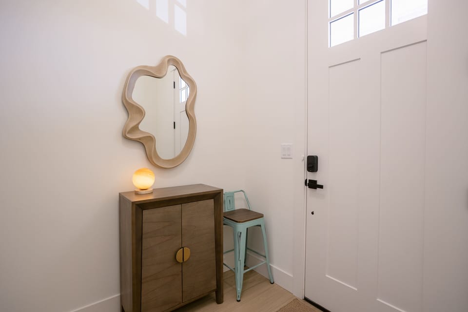 A welcoming entry space with a uniquely shaped mirror, a small wooden cabinet, a soft glowing lamp, and a teal accent stool by a modern white front door with smart lock access.