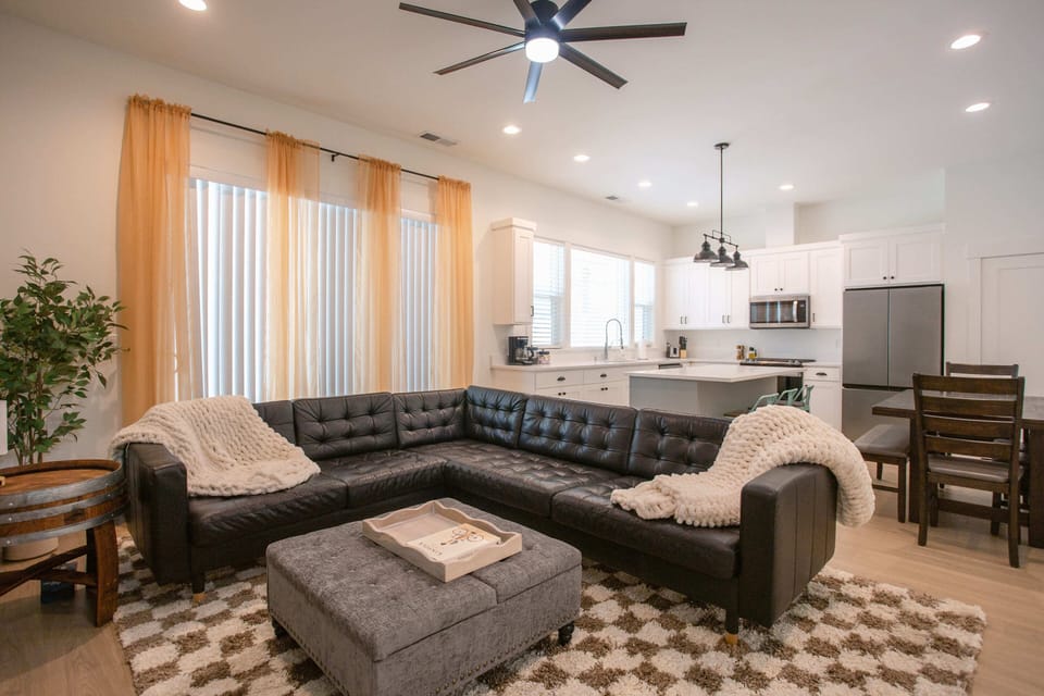 A stylish open-concept living area featuring a black leather sectional sofa with cozy throws, a patterned rug, and a modern kitchen with white cabinetry, stainless steel appliances, and bar seating.