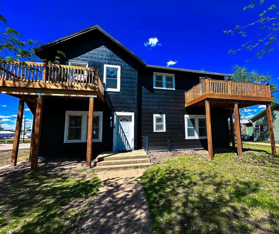 This striking house features bold black vinyl siding and elevated wooden decks, perfect for outdoor relaxation. Its unique design offers a modern aesthetic, complemented by a well-maintained lawn and inviting entryway.