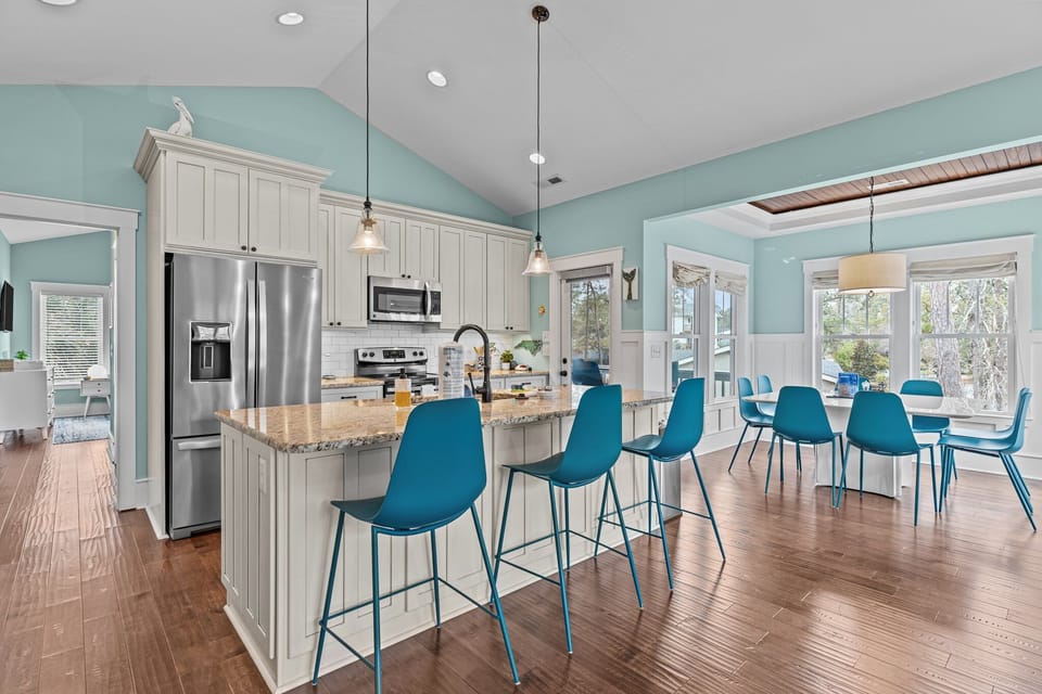 Bright kitchen, granite island, teal stools, white cabinets, stainless fridge, pendant lights, wood floors, dining table, windows, vaulted ceiling.