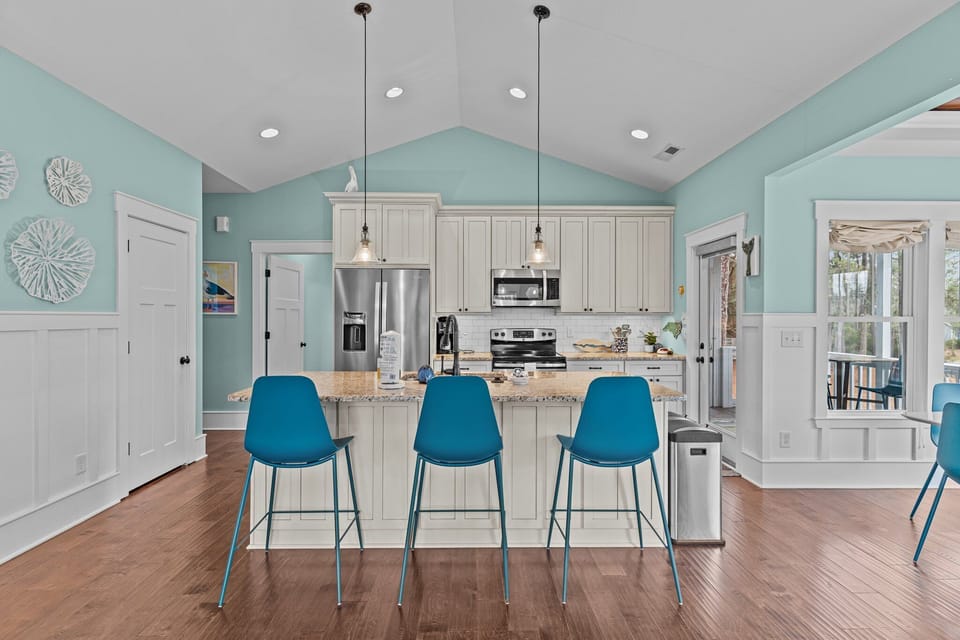 Open kitchen living area, granite island sink, pendant lights, teal chairs, sofa, wall TV, large windows, ceiling fan, beach decor, wood floors.