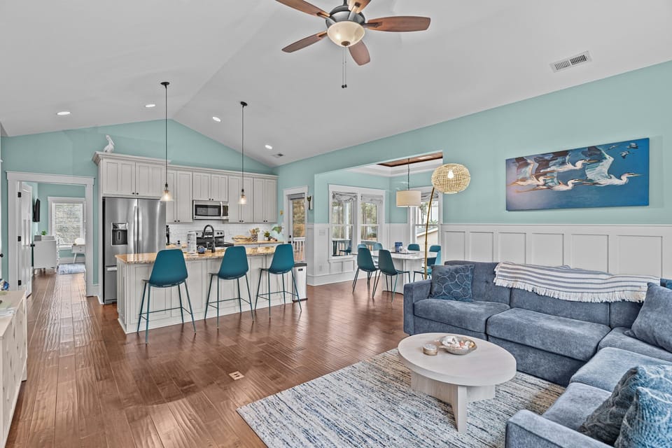 Open kitchen living area, granite island sink, pendant lights, teal chairs, sofa, wall TV, large windows, ceiling fan, beach decor, wood floors.