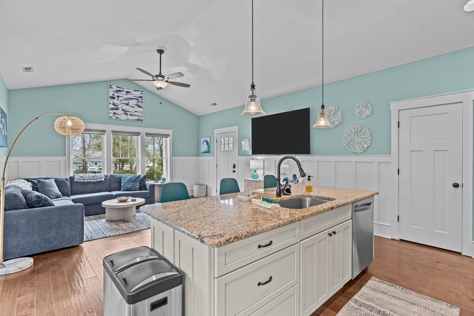 Open kitchen living area, granite island sink, pendant lights, teal chairs, sofa, wall TV, large windows, ceiling fan, beach decor, wood floors.