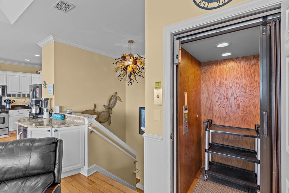Oak Island NC vacation rental interior showing a private elevator with wood-paneled interior, adjacent stairwell, coastal wall decor, wood floors, and open access to the kitchen and living area in a beach house.