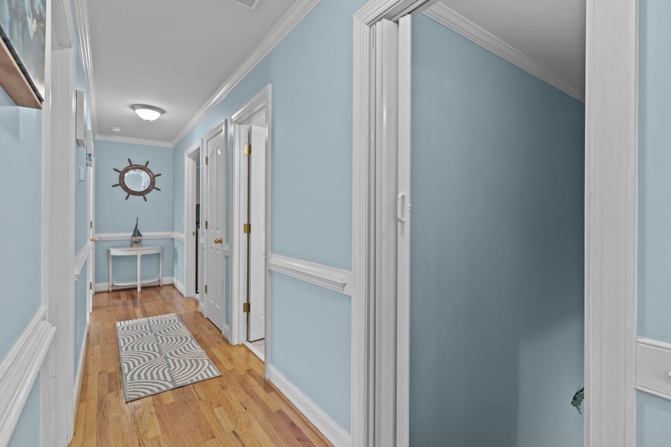 Light blue hallway featuring hardwood floors, white trim, multiple bedroom doors, runner rug, and coastal accents including ship wheel mirror and decorative table.