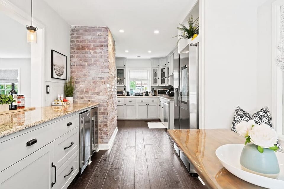 Modern kitchen features white cabinetry, a brick accent wall, stainless steel appliances, and a wooden dining table.
