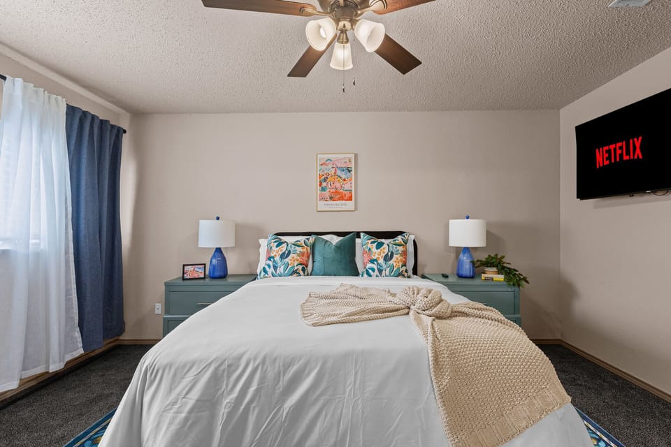 Serene bedroom with a white bed, teal headboard, and orange pillows, complemented by a green dresser and ceiling fan.