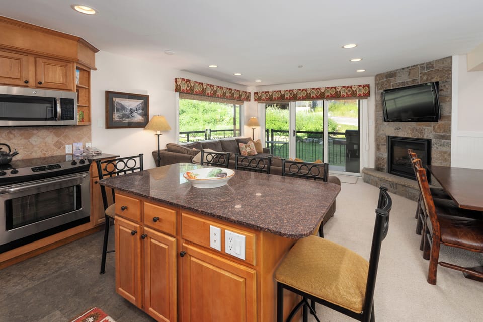 Inviting open-concept living space featuring a modern kitchen flowing seamlessly into a cozy living area with a sectional sofa, stone fireplace, and large windows that open to a wrap around patio.