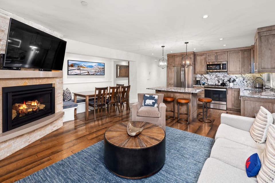 Inviting open-concept living space featuring a modern kitchen flowing seamlessly into a cozy living area with a sectional sofa, stone fireplace, and large windows that open to a wrap around balcony.