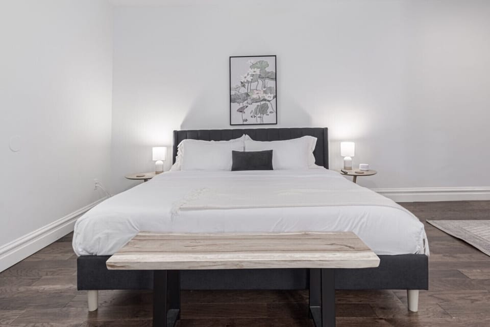 Primary Bedroom Suite: Unwind in ultimate comfort after a day of exploring, featuring a king bed with premium linens, upholstered headboard, bedside tables with reading lamps, and dark hardwood floors in a spacious, minimalist setup.