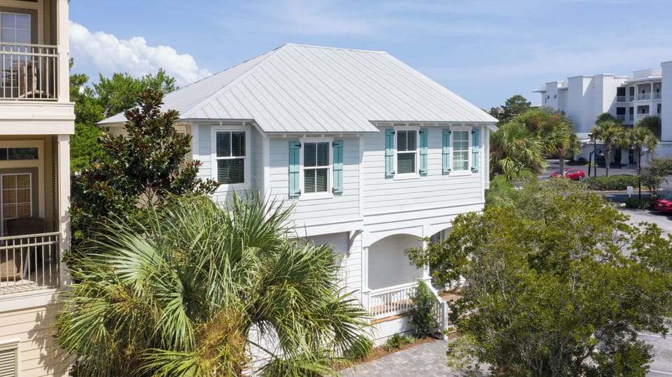 Bright coastal exterior with white siding, metal roof, and charming aqua shutters—nestled among palms for a relaxed beach-town feel.