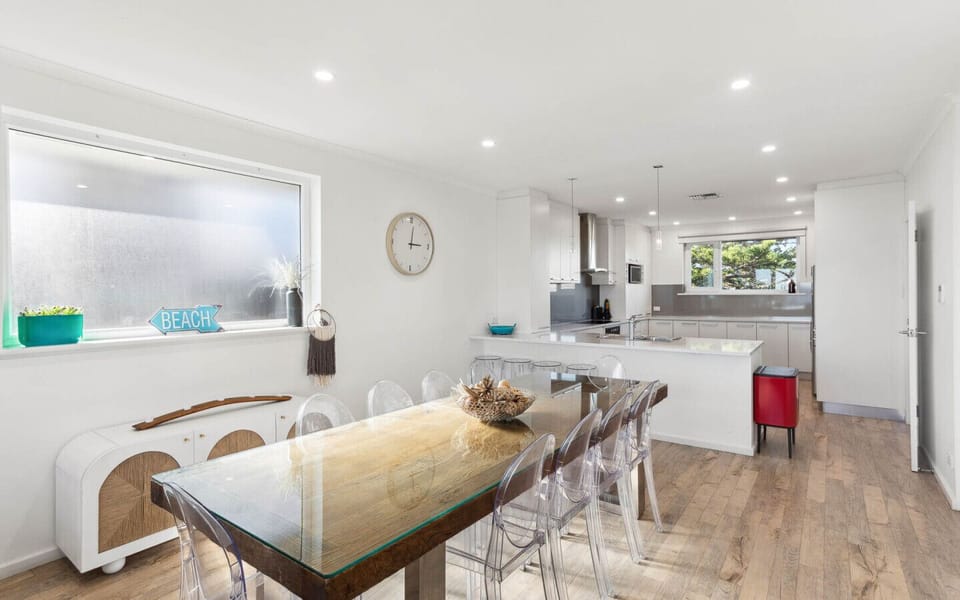 The space features a wooden dining table with transparent chairs, a wall clock, a `BEACH` sign on the window sill, and a red trash bin near the kitchen area, which has a sleek design with stainless steel appliances. The overall look is bright and inviting