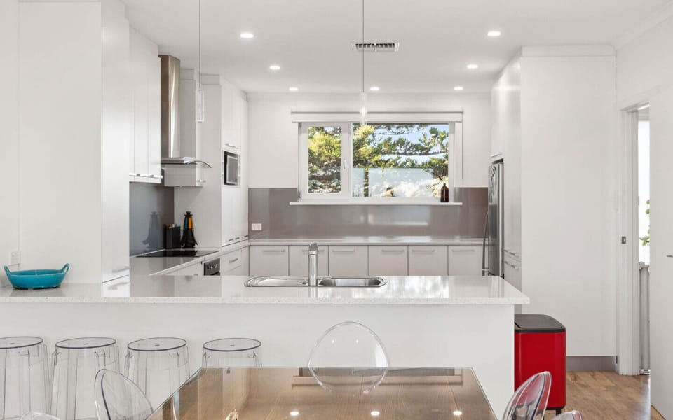 A modern, bright kitchen with a predominantly white color scheme. It features sleek cabinetry, a central island with a countertop, and a large window that allows natural light to flood the space. The kitchen is equipped with stainless steel appliances, an
