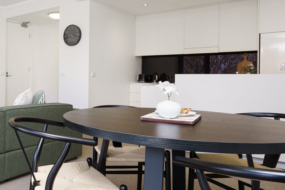 Simple and modern designed dining area with a dark round table and comfortable chairs, complemented by a touch of greenery and large windows that bring in plenty of natural light.