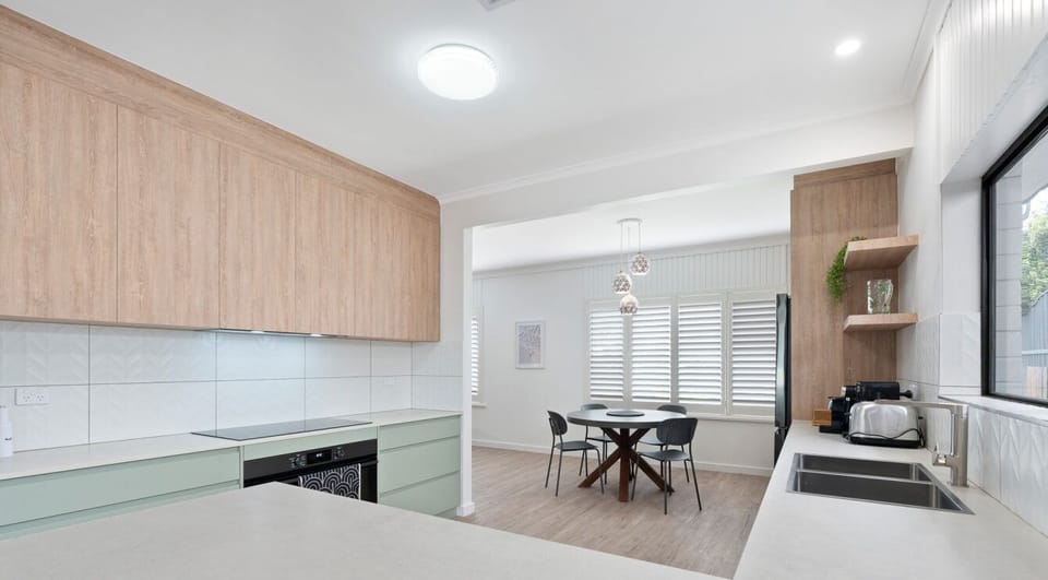 A modern kitchen with light-colored countertops and a combination of wood and pastel green cabinetry. There is a dining area in the background with a table and chairs, and the space features large windows with shutters, allowing natural light to flood in.