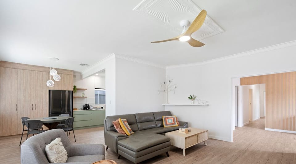 A modern, stylish living space with a light color scheme. It features a gray sofa with colorful cushions, matching gray armchairs, and a wooden coffee table. The area has wooden flooring and includes a ceiling fan, pendant lights, and wooden cabinetry in 