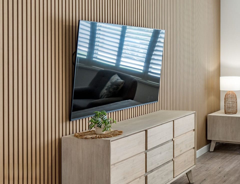 A modern interior space with a large TV mounted on a wall with vertical wooden paneling. Below the TV is a light wooden dresser with a small plant on top. To the right, there is a side table with a woven lamp, contributing to a cozy and stylish atmosphere