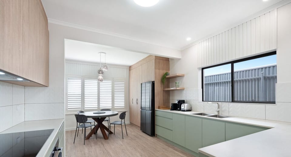 A modern kitchen with light green cabinets and a light wood floor. It features a dining area with a round table and chairs, large windows with shutters for natural light, and a sleek design with integrated appliances and a clean, minimalist aesthetic.