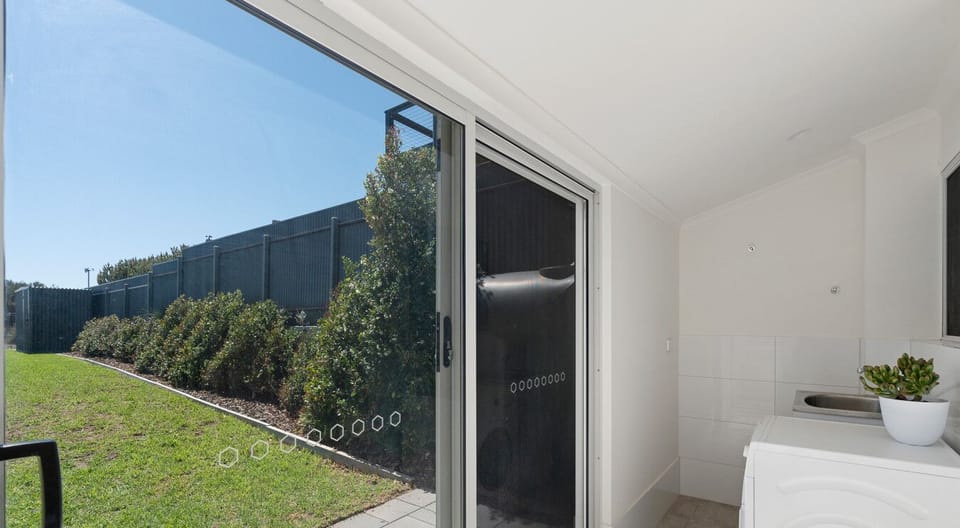 A room with a large sliding door that opens to a view of a grassy area with bushes and a fenced background. Inside the room, there is a white appliance, possibly a washing machine, with a potted plant on top, and the space has light-colored walls and tile