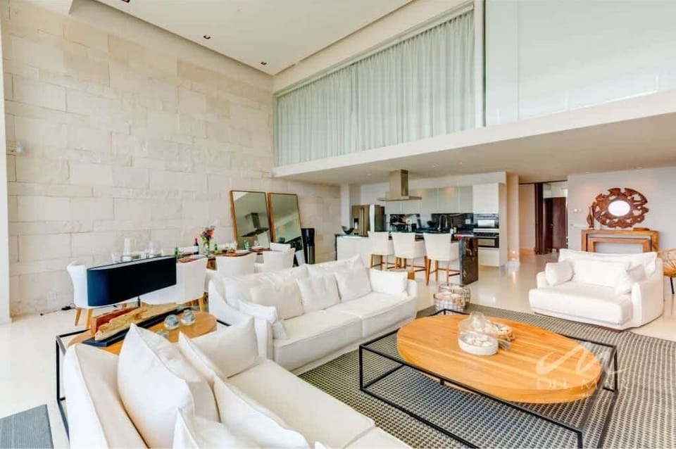 Bright living room w/ white sofas, wood accents & floor-to-ceiling windows inviting in soft daylight