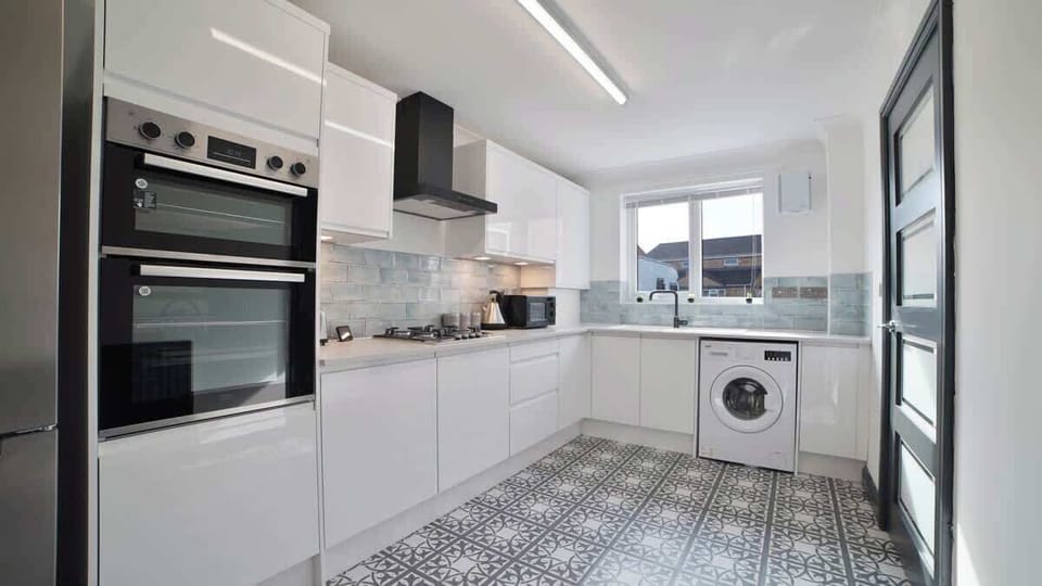 Sleek and modern full kitchen featuring glossy white cabinets, built-in double ovens, a gas cooktop, washing machine, and patterned tile flooring—designed for both style and functionality.
