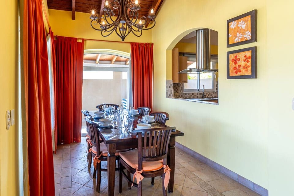 Cozy dining area with a wooden table for six, complemented by vibrant yellow walls and bold red curtains. Features a chandelier and a seamless view of the kitchen, creating an inviting atmosphere for meals and gatherings.