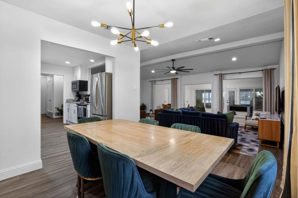 Elegant open-concept dining room with a large solid-wood 6-seat table, modern gold sputnik chandelier, emerald velvet chairs, and open views into the kitchen and living room — the perfect setting for big family dinners and celebrations.