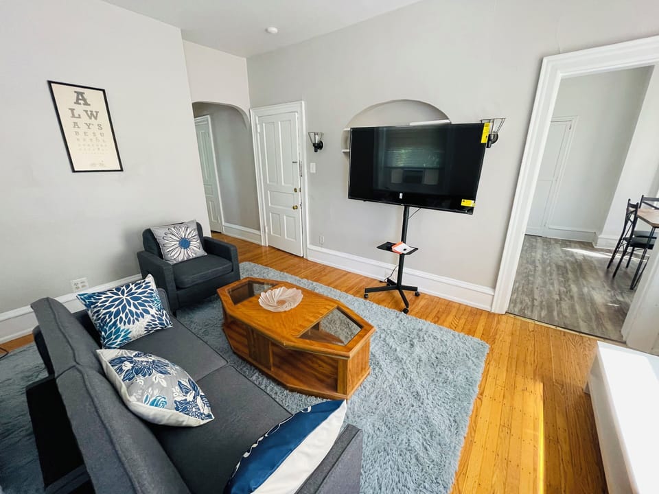 Cozy, sunlit living room with hardwood floors, sectional sofa, accent chairs, and a modern coffee table. Features built-in shelving, mounted TV, and stylish decor—perfect for relaxing or entertaining in comfort.