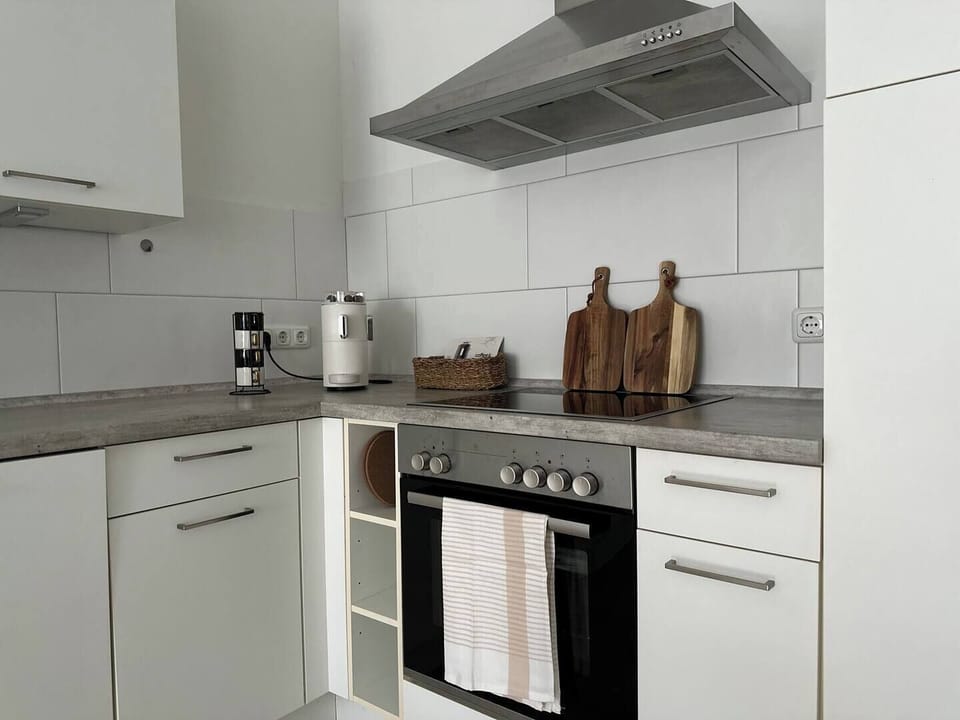 Moderne Küchenzeile mit Cerankochfeld, Backofen und Dunstabzugshaube; praktische Arbeitsfläche und Zubehör wie Schneidebretter, Wasserkocher und Kapselmaschine.